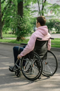 Person in a pink hoodie sitting in a wheelchair along a park path facing away from the camera