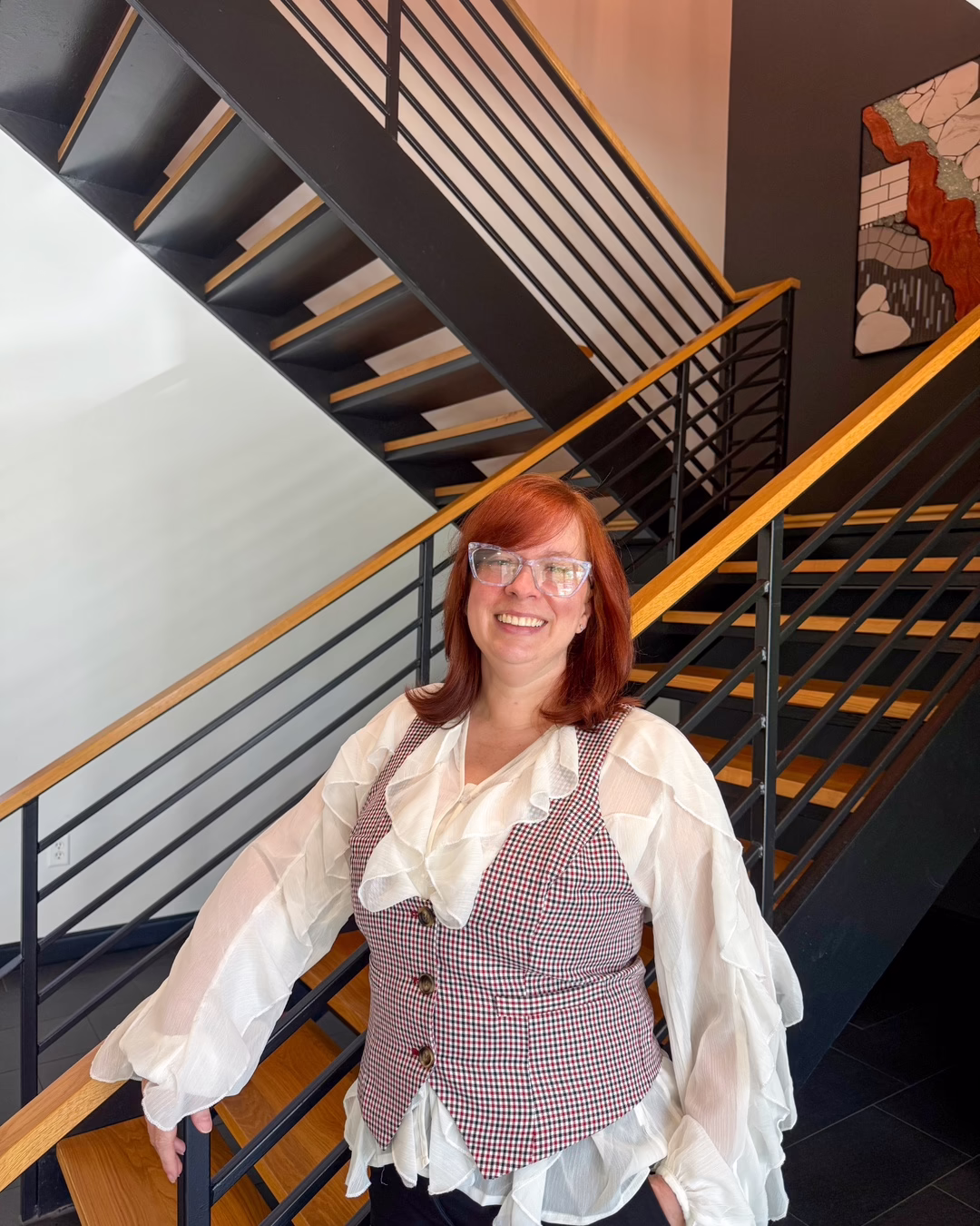 Smiling woman with red hair wearing a white ruffled blouse and checkered vest standing on a modern wooden staircase indoors