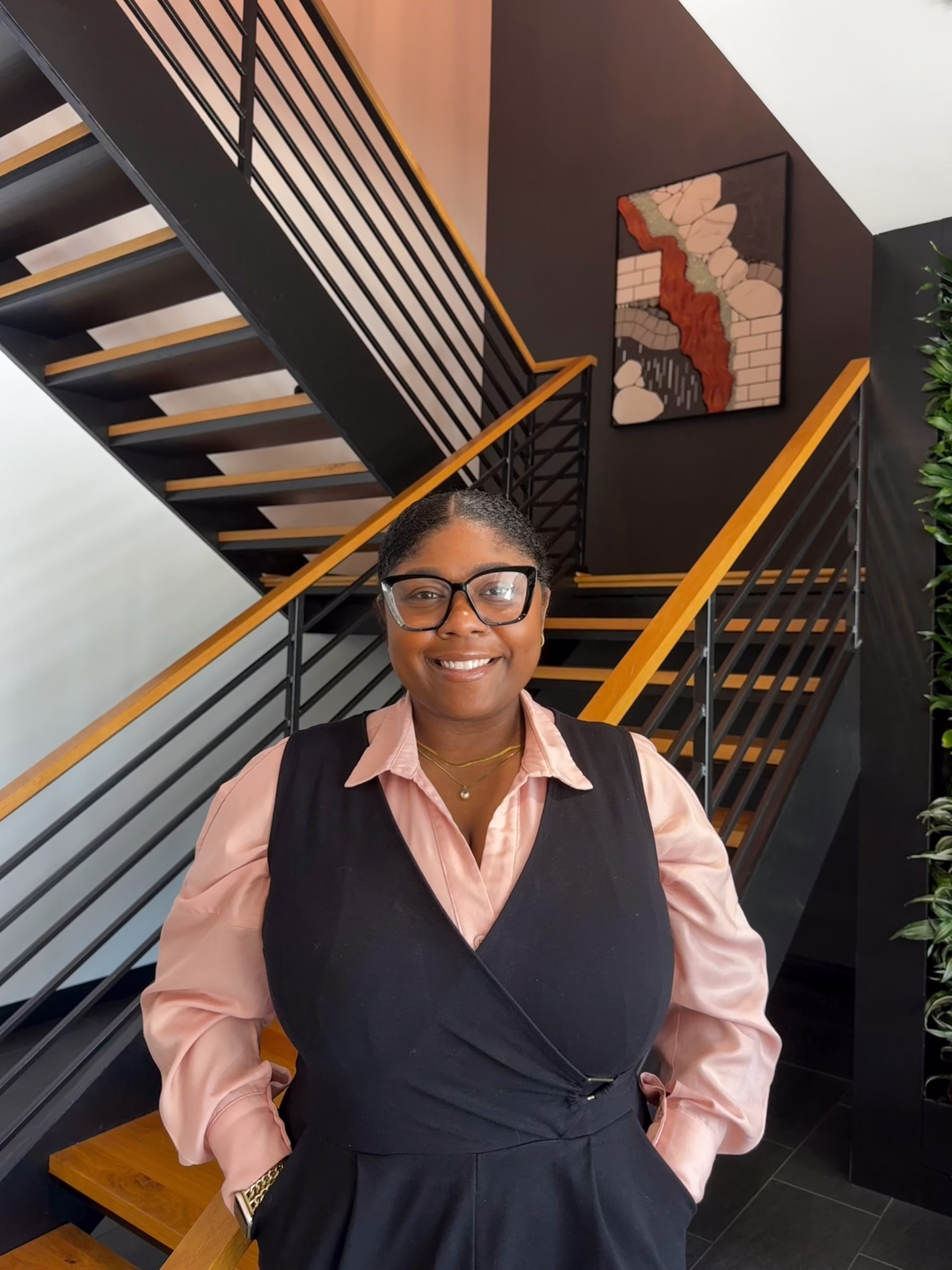 Smiling woman in a pink blouse and black vest stands beside a modern staircase with wood and metal railings in a stylish interior